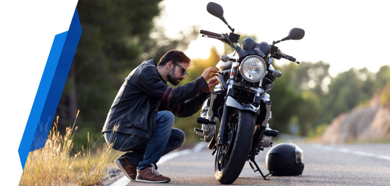 A man kneeling beside a motorcycle on the roadside, inspecting it with his helmet placed nearby, waiting for Smartcover Motorcycle Breakdown roadside assistance.